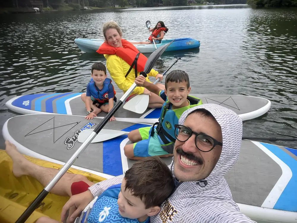 Danny on a paddleboard taking a selfie on a lake, with a woman and four children in life vests on boards and a kayak