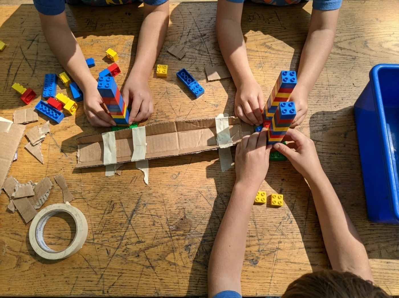 Two pairs of children's hands building LEGO towers on a scratched wooden table with cardboard and tape