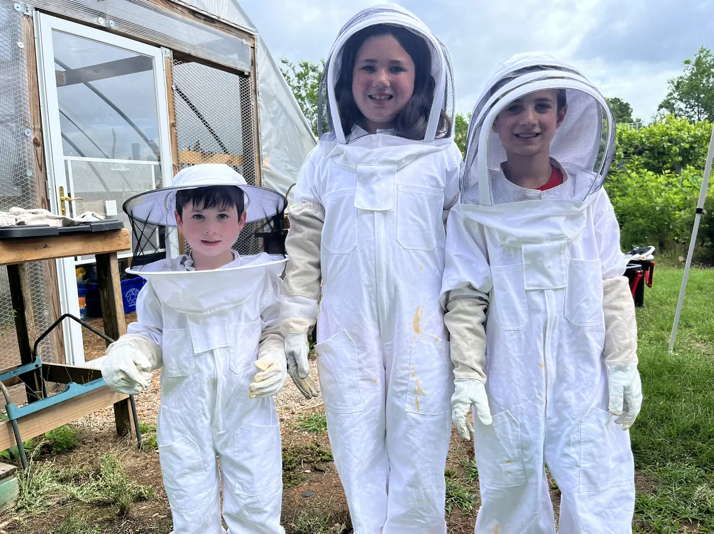 Three campers in full white bee suits and mesh veils, smiling shoulder to shoulder outside a hoop greenhouse